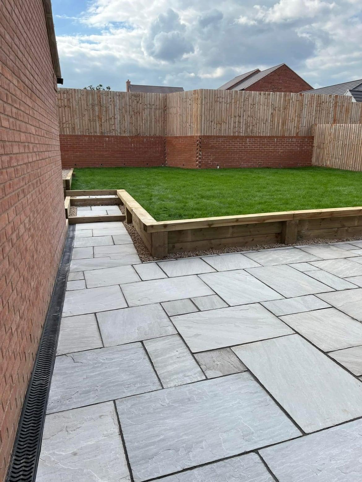 Light sandstone patio with a drainage channel, timber sleeper retaining edge and a fresh lawn