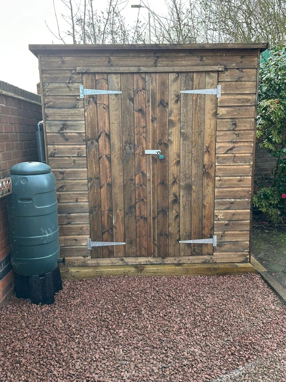 Timber shed with double doors on a red gravel base, with a water butt alongside
