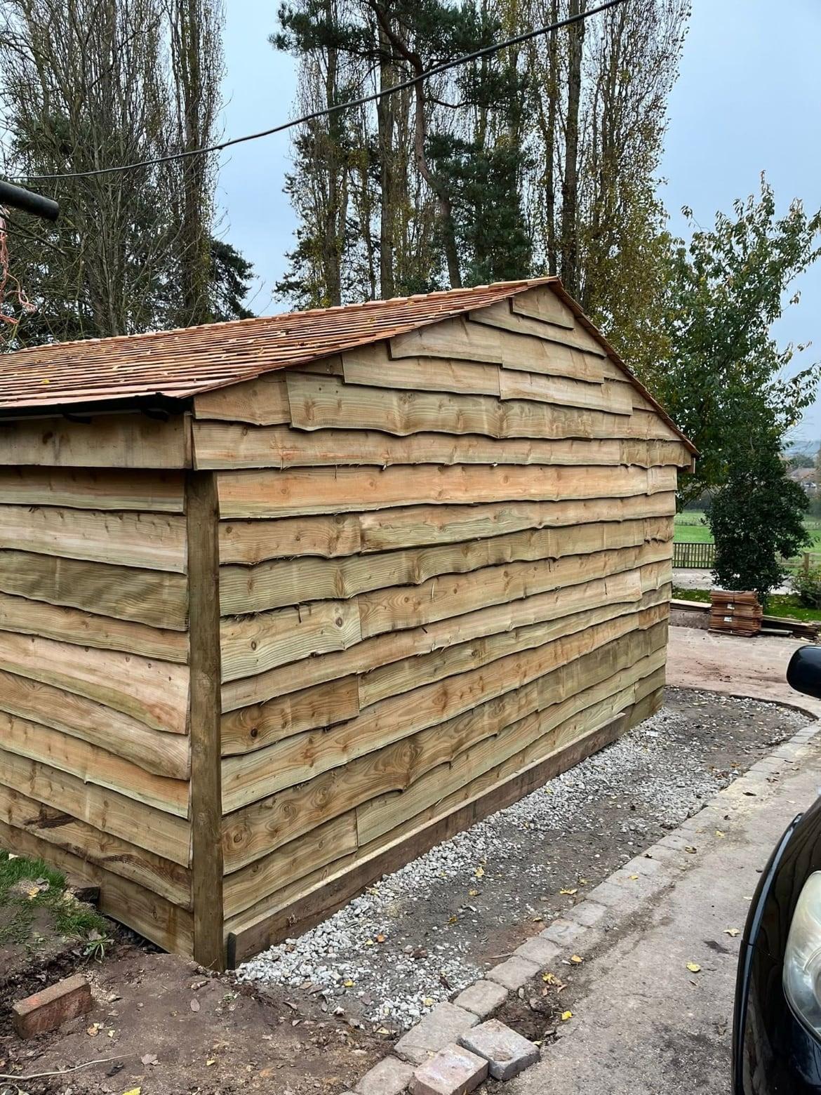 Waney-edge timber garage with a tiled roof beside a gravel path